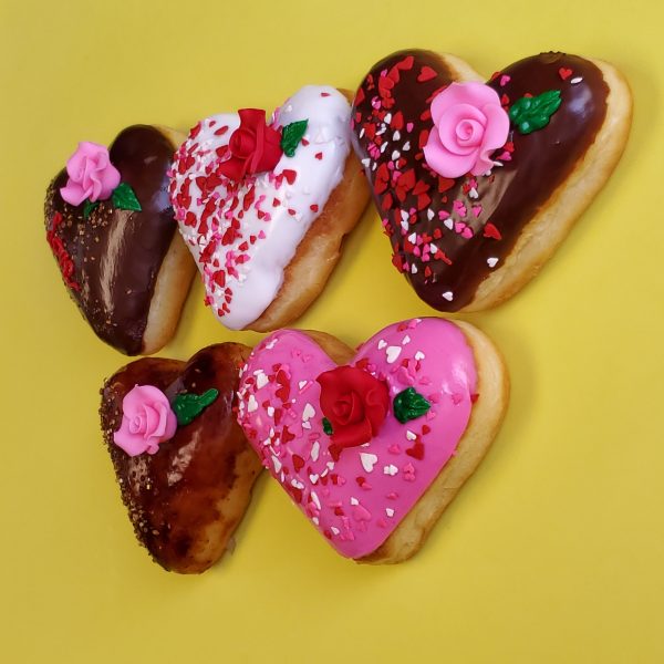 A beautifully decorated Valentine’s Pink Rose Heart Donut, coated in smooth pink icing, sprinkled with heart-shaped sprinkles, gold star accents, and edible gold sprinkles, and topped with a delicate candy rose. A perfect romantic treat for Valentine’s Day.