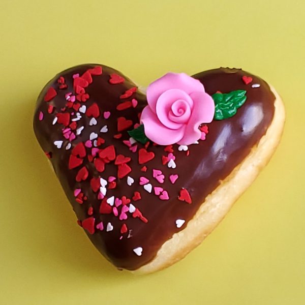 A beautifully decorated Valentine’s Chocolate Rose Heart Donut, coated in smooth chocolate icing, sprinkled with gold star sprinkles, heart-shaped decorations, and edible gold accents, and topped with a delicate candy rose. A perfect romantic treat for Valentine’s Day.