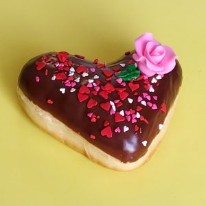 A beautifully decorated Valentine’s Chocolate Rose Heart Donut, coated in smooth chocolate icing, sprinkled with gold star sprinkles, heart-shaped decorations, and edible gold accents, and topped with a delicate candy rose. A perfect romantic treat for Valentine’s Day