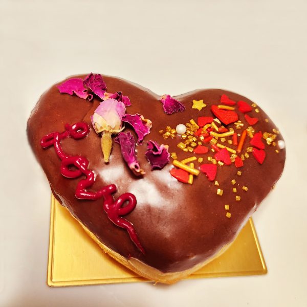 A beautifully decorated Valentine’s Love Heart Donut, filled with strawberry jam, topped with a smooth chocolate glaze, adorned with gold sprinkles, edible star accents, and a delicate candy rose, featuring a hand-piped ‘Love’ design. A perfect romantic treat for Valentine’s Day.