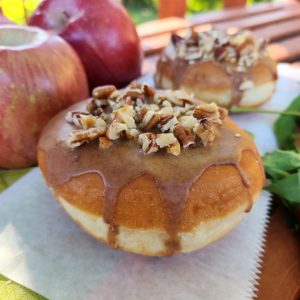 A Thanksgiving donut with a round éclair shape, filled with purple yam jam, topped with a cinnamon glaze and a mix of chopped nuts. The donut is displayed with fresh apples in the background for a seasonal touch.