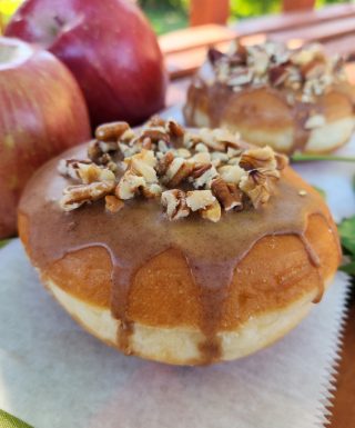 A Thanksgiving donut with a round éclair shape, filled with purple yam jam, topped with a cinnamon glaze and a mix of chopped nuts. The donut is displayed with fresh apples in the background for a seasonal touch.