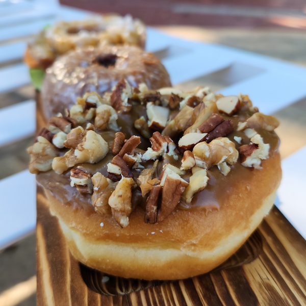 A Thanksgiving donut with a maple icing glaze, topped with a mix of chopped pecans, walnuts, and almonds. The donut is fluffy and maple glaze with crunchy nut topping that adds texture and flavor. Perfect for the holiday season.