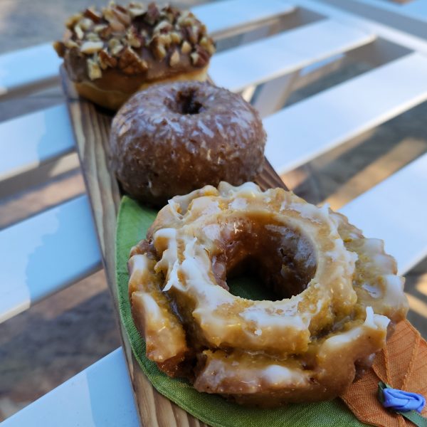 Thanksgiving donuts displayed, including a Pumpkin Buttermilk donut with a light glaze, surrounded by a Maple Icing with Mixed Nuts donut and a classic glazed donut.