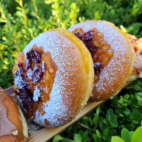 A Thanksgiving donut with sweet orange marmalade and cranberry topping, lightly dusted with powdered sugar. The round éclair-style donut is filled with orange jam and decorated with fresh fruits in the background.