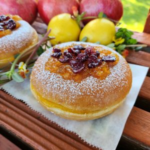 A Thanksgiving donut with sweet orange marmalade and cranberry topping, lightly dusted with powdered sugar. The round éclair-style donut is filled with orange jam and decorated with fresh fruits in the background.