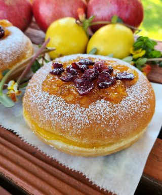 A Thanksgiving donut with sweet orange marmalade and cranberry topping, lightly dusted with powdered sugar. The round éclair-style donut is filled with orange jam and decorated with fresh fruits in the background.