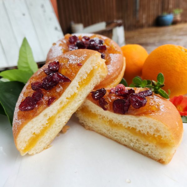 A Thanksgiving donut sliced in half to reveal a filling of smooth orange jam. The donut is topped with sweet orange marmalade and dried cranberries, with a light dusting of powdered sugar. Fresh oranges and green leaves decorate the background, enhancing the festive presentation.