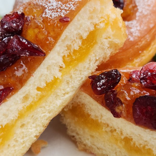 A Thanksgiving donut sliced in half to reveal a filling of smooth orange jam. The donut is topped with sweet orange marmalade and dried cranberries, with a light dusting of powdered sugar. Fresh oranges and green leaves decorate the background, enhancing the festive presentation.