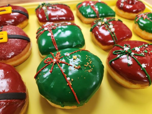 A round donut with green icing, decorated with a red ribbon design and festive sprinkles including gold stars, pearls, and snowflakes.