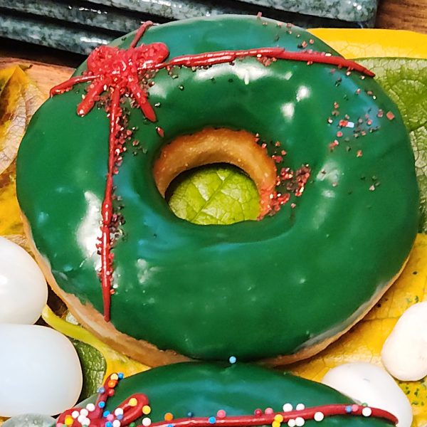 A Christmas-themed donut with green icing, decorated with red ribbon details and colorful sprinkles, perfect for holiday celebrations.