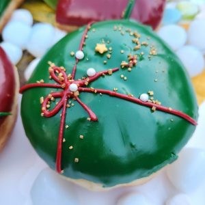 A round donut with green icing, decorated with a red ribbon design and festive sprinkles including gold stars, pearls, and snowflakes.