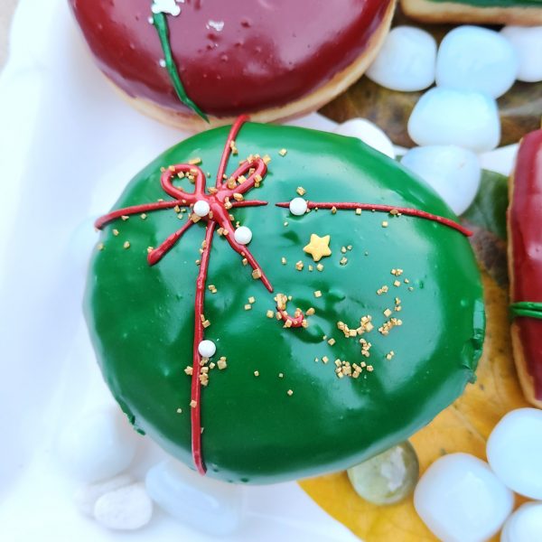 A round donut with green icing, decorated with a red ribbon design and festive sprinkles including gold stars, pearls, and snowflakes.