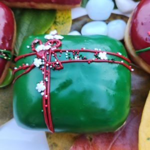 A square donut with green icing, decorated with a red ribbon design and festive sprinkles including gold stars, pearls, and snowflakes.