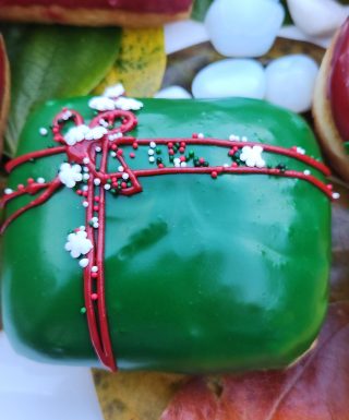 A square donut with green icing, decorated with a red ribbon design and festive sprinkles including gold stars, pearls, and snowflakes.