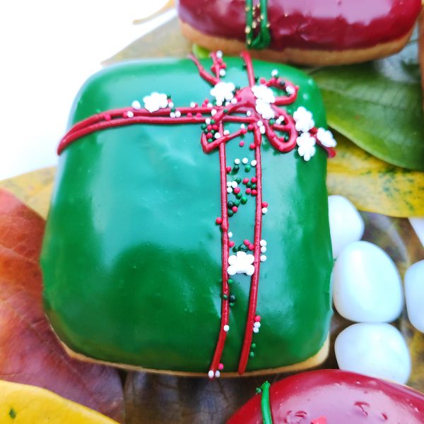 A square donut with green icing, decorated with a red ribbon design and festive sprinkles including gold stars, pearls, and snowflakes.