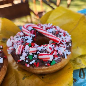 A donut with chocolate icing, topped with crushed candy canes, festive sprinkles, snowflake accents, and a whole candy cane, perfect for Christmas celebrations.