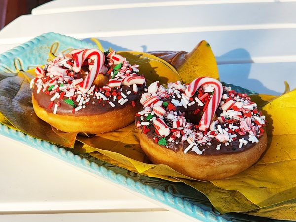 A donut with chocolate icing, topped with crushed candy canes, festive sprinkles, snowflake accents, and a whole candy cane, perfect for Christmas celebrations.