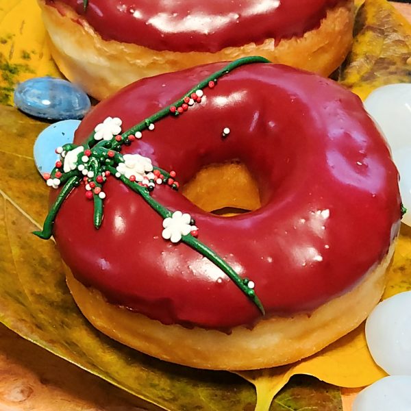 A Christmas-themed donut with red icing, decorated with green ribbon details and colorful sprinkles, perfect for holiday celebrations.