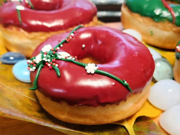 A Christmas-themed donut with red icing, decorated with green ribbon details and colorful sprinkles, perfect for holiday celebrations.