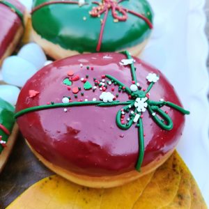 A round donut with red icing, decorated with a green ribbon design and festive sprinkles including gold stars, pearls, and snowflakes.