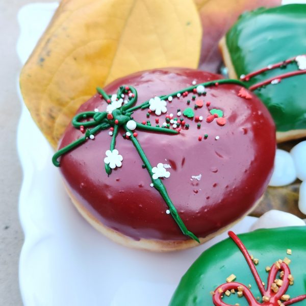 A round donut with red icing, decorated with a green ribbon design and festive sprinkles including gold stars, pearls, and snowflakes.