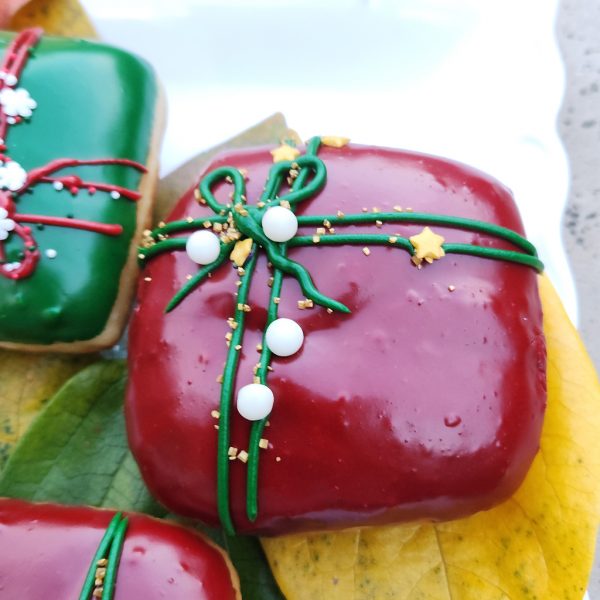 A square donut with red icing, decorated with a green ribbon design and festive sprinkles including gold stars, pearls, and snowflakes.