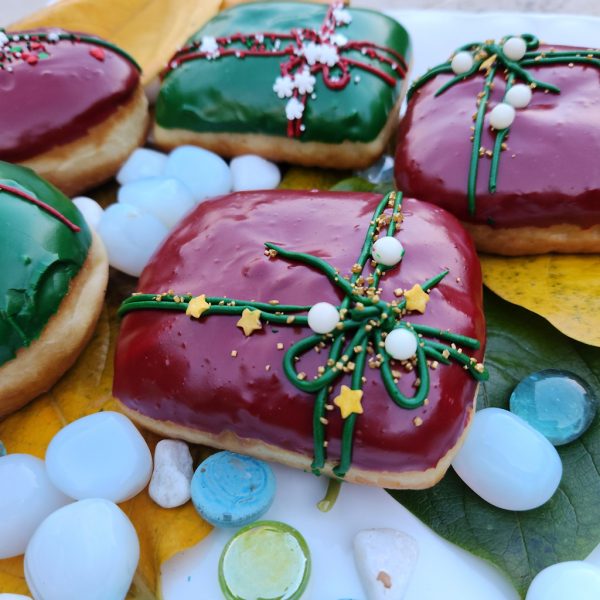 A square donut with red icing, decorated with a green ribbon design and festive sprinkles including gold stars, pearls, and snowflakes.