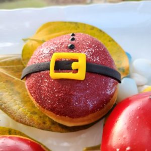 A round Christmas donut with red icing and sprinkle sugar, decorated with a candy black belt and yellow buckle, resembling Santa Claus's outfit.