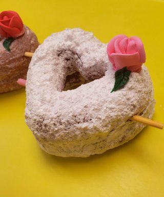 A beautifully crafted Valentine’s Powdered Sugar Croissant Cupid Donut, dusted with fine powdered sugar, topped with a delicate candy rose, and pierced with a strawberry cream Pocky stick as Cupid’s arrow. A crispy, flaky croissant donut perfect for Valentine’s Day.