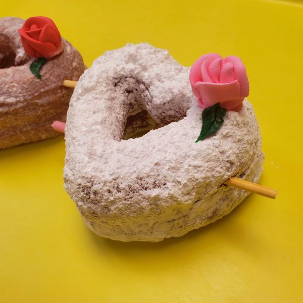 A beautifully crafted Valentine’s Powdered Sugar Croissant Cupid Donut, dusted with fine powdered sugar, topped with a delicate candy rose, and pierced with a strawberry cream Pocky stick as Cupid’s arrow. A crispy, flaky croissant donut perfect for Valentine’s Day.