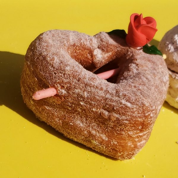 A beautifully crafted Valentine’s Croissant Cupid Heart Donut, dusted with fine sugar, topped with a delicate candy rose, and pierced with a strawberry cream Pocky stick as Cupid’s arrow. A crispy, flaky croissant donut perfect for Valentine’s Day.