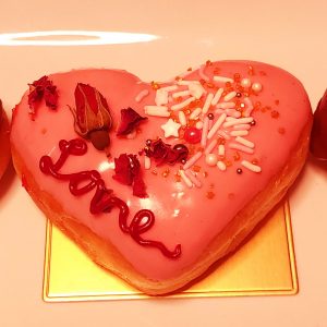 A beautifully decorated Valentine’s Pink Love Heart Donut, filled with strawberry jam, coated in smooth pink icing, adorned with gold sprinkles, edible dried rose petals, candy stars, and a piped ‘Love’ design. A perfect romantic treat for Valentine’s Day.