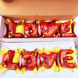 A beautifully crafted Valentine’s LOVE Letter Donut Set, coated in a shiny strawberry mirror glaze, decorated with edible gold flakes and dried rose petals, placed in an elegant gold tray and presented in a Valentine’s-themed gift box. A perfect romantic dessert for gifting.