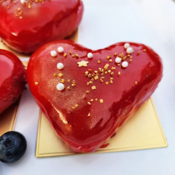 A beautifully crafted Valentine’s Heart Donut, filled with rich strawberry jam, coated in a glossy strawberry mirror glaze, decorated with gold sprinkles, pearl sugar, and edible star accents, placed on an elegant gold tray with fresh blueberries on the side. A sweet and romantic treat for Valentine’s Day.