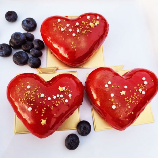 A beautifully crafted Valentine’s Heart Donut, coated in a glossy strawberry mirror glaze, decorated with gold sprinkles, pearl sugar, and edible star accents, placed on an elegant gold tray with fresh blueberries on the side. A sweet and romantic treat for Valentine’s Day