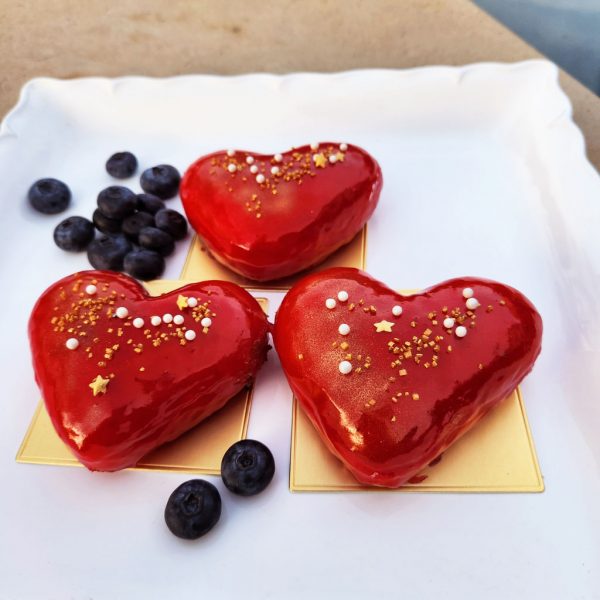 A beautifully crafted Valentine’s Heart Donut, coated in a glossy strawberry mirror glaze, decorated with gold sprinkles, pearl sugar, and edible star accents, placed on an elegant gold tray with fresh blueberries on the side. A sweet and romantic treat for Valentine’s Day.