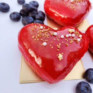 A beautifully crafted Valentine’s Heart Donut, filled with rich strawberry jam, coated in a glossy strawberry mirror glaze, decorated with gold sprinkles, pearl sugar, and edible star accents, placed on an elegant gold tray with fresh blueberries on the side. A sweet and romantic treat for Valentine’s Day.