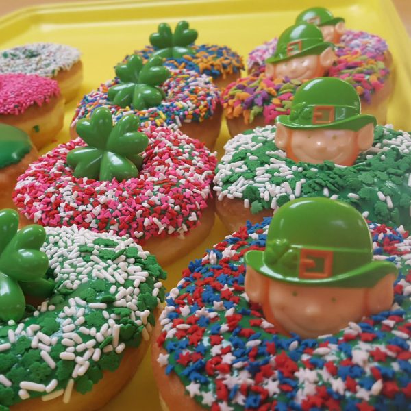 A St. Patrick’s Day-themed donut with green, white, and gold sprinkles, topped with a fun holiday ring featuring shamrocks, leprechauns, and gold coin designs.