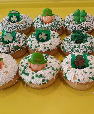 A St. Patrick’s Day-themed donut with green, white, and gold sprinkles, topped with a fun holiday ring featuring shamrocks, leprechauns, and gold coin designs.