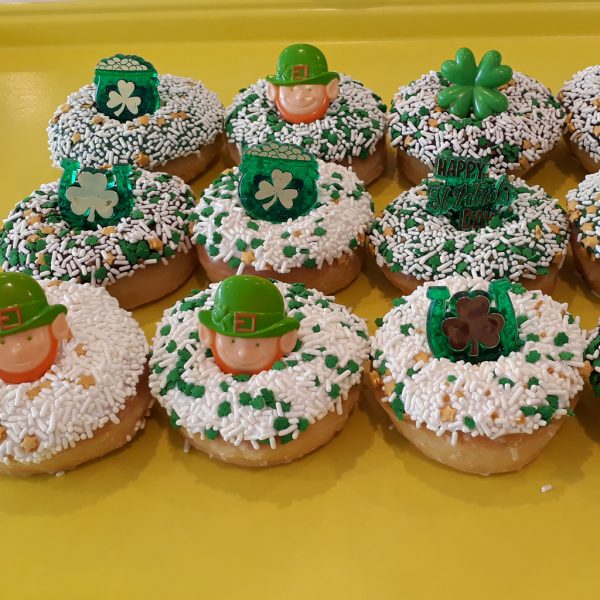 A St. Patrick’s Day-themed donut with green, white, and gold sprinkles, topped with a fun holiday ring featuring shamrocks, leprechauns, and gold coin designs.