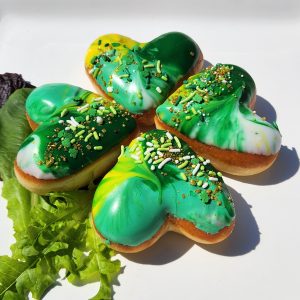 A set of four heart-shaped donuts with green, white, and yellow galaxy icing, arranged to form a lucky four-leaf clover, topped with festive sprinkles and gold shimmer.