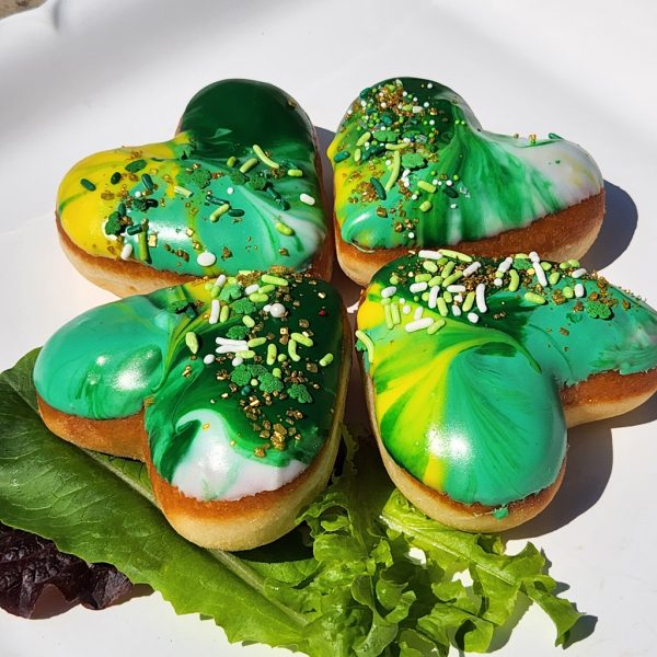 A set of four heart-shaped donuts with green, white, and yellow galaxy icing, arranged to form a lucky four-leaf clover, topped with festive sprinkles and gold shimmer.