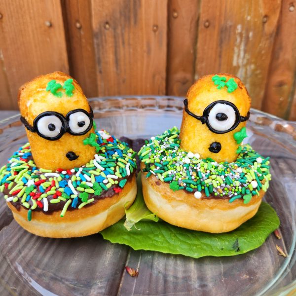 A St. Patrick’s Day-themed donut covered in green, white, and gold sprinkles, topped with a Twinkie Minion character decorated with a lucky clover design.