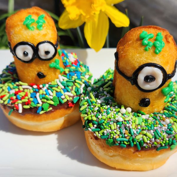 A St. Patrick’s Day-themed donut covered in green, white, and gold sprinkles, topped with a Twinkie Minion character decorated with a lucky clover design.