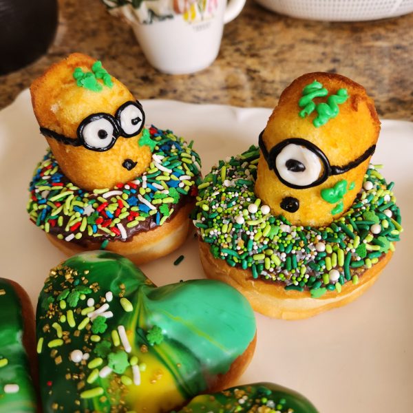 A St. Patrick’s Day-themed donut covered in green, white, and gold sprinkles, topped with a Twinkie Minion character decorated with a lucky clover design.