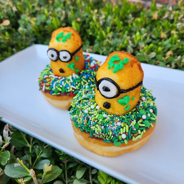 A St. Patrick’s Day-themed donut covered in green, white, and gold sprinkles, topped with a Twinkie Minion character decorated with a lucky clover design.