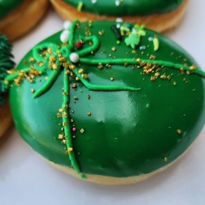 A round donut covered in smooth green icing, decorated with a piped green ribbon, gold sprinkles, and festive shamrock accents for St. Patrick’s Day.