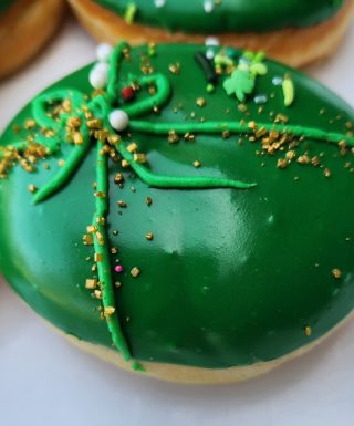 A round donut covered in smooth green icing, decorated with a piped green ribbon, gold sprinkles, and festive shamrock accents for St. Patrick’s Day.