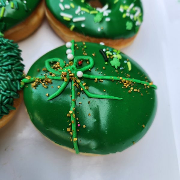 A round donut covered in smooth green icing, decorated with a piped green ribbon, gold sprinkles, and festive shamrock accents for St. Patrick’s Day.